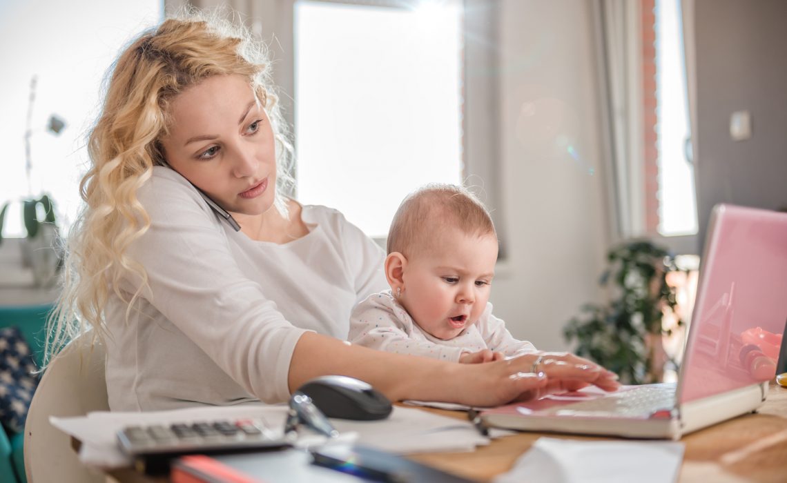 Mother holding baby, talking on smart phone at home office and using laptop