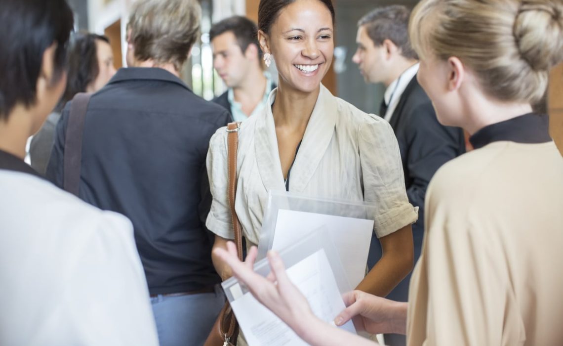 Portrait of two smiling women holding files and talking, standing in crowded lobby